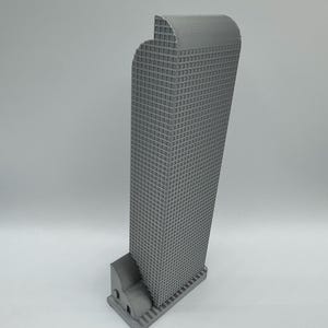 May include: A gray, 3D-printed model of a skyscraper. The building has a rectangular base with a curved top and a grid-like pattern on the facade, resembling windows. The model is set against a plain, light gray background.