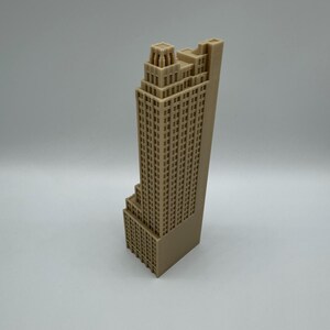 May include: A tan-colored, 3D-printed model of a skyscraper. The building has a stepped design with many windows and architectural details. The model is standing upright against a plain background.
