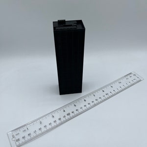 May include: A black miniature building model with a flat roof and a ruler for scale. The building is approximately 3 inches tall.
