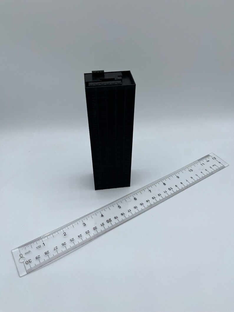 May include: A black miniature building model with a flat roof and a ruler for scale. The building is approximately 3 inches tall.