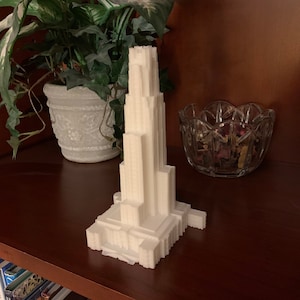 May include: A white 3D printed model of a tall building with a square base and many windows. The model is sitting on a wooden shelf.