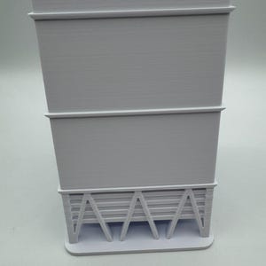 May include: A light gray, 3D-printed architectural model. The model features a rectangular base supporting a tiered structure with horizontal lines and angled supports, showcasing a minimalist design.