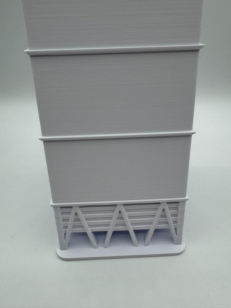 May include: A light gray, 3D-printed architectural model. The model features a rectangular base supporting a tiered structure with horizontal lines and angled supports, showcasing a minimalist design.
