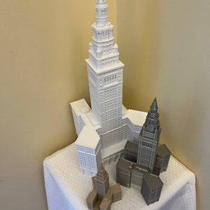 May include: Three white and gray 3D printed models of a skyscraper. The tallest model is white and the two smaller models are gray. The models are sitting on a white tablecloth.
