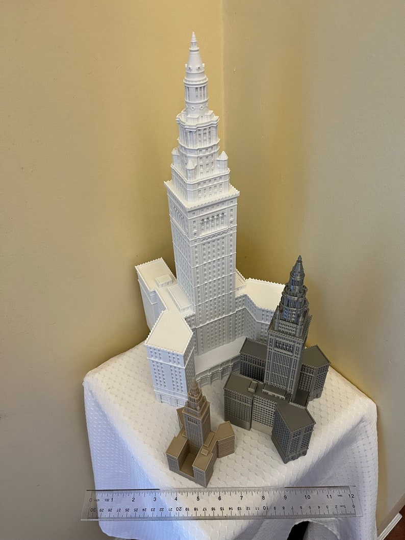 May include: Three white and gray 3D printed models of a skyscraper. The tallest model is white and the two smaller models are gray. The models are sitting on a white tablecloth.