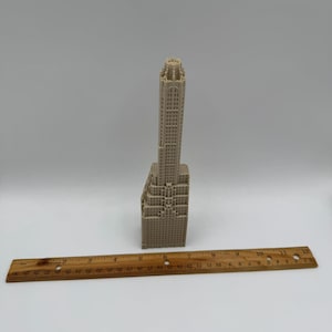 May include: A beige miniature skyscraper model with intricate architectural details. A wooden ruler is placed horizontally below the building. The model is a detailed representation of a tall building.