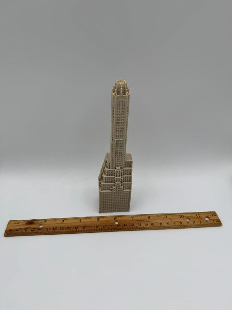 May include: A beige miniature skyscraper model with intricate architectural details. A wooden ruler is placed horizontally below the building. The model is a detailed representation of a tall building.