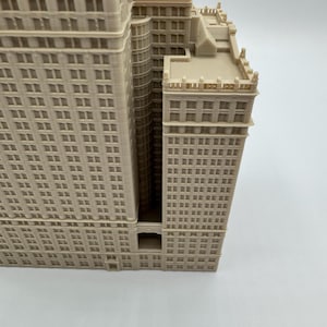 May include: A beige architectural model of a multi-story building with numerous windows. The building has a detailed facade and a recessed central section. The model is a detailed representation of a skyscraper.