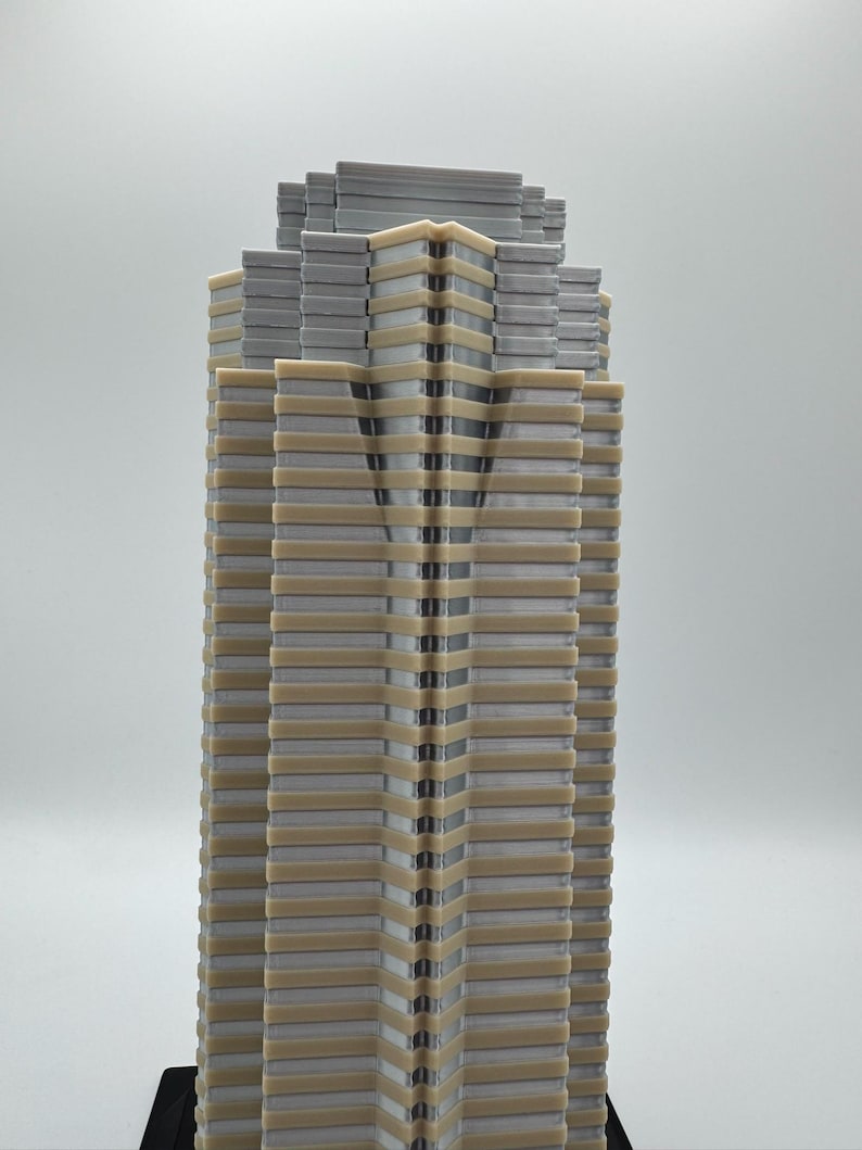 May include: A detailed model of a skyscraper, featuring a layered design with alternating gray and beige sections. The building's facade has a vertical pattern, and the top section has a stepped appearance. The model is set against a plain background.
