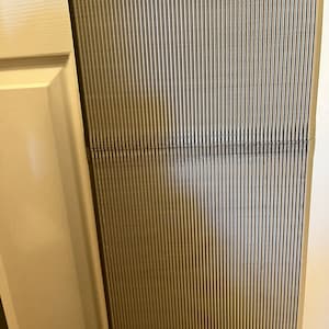 Pu&ograve; includere: Una sezione di una superficie verticale e scanalata in tonalit&agrave; di grigio e beige. Le scanalature sono parallele, creando un aspetto strutturato. Una linea orizzontale divide la superficie. Il bordo di un telaio di porta bianco &egrave; visibile a sinistra.