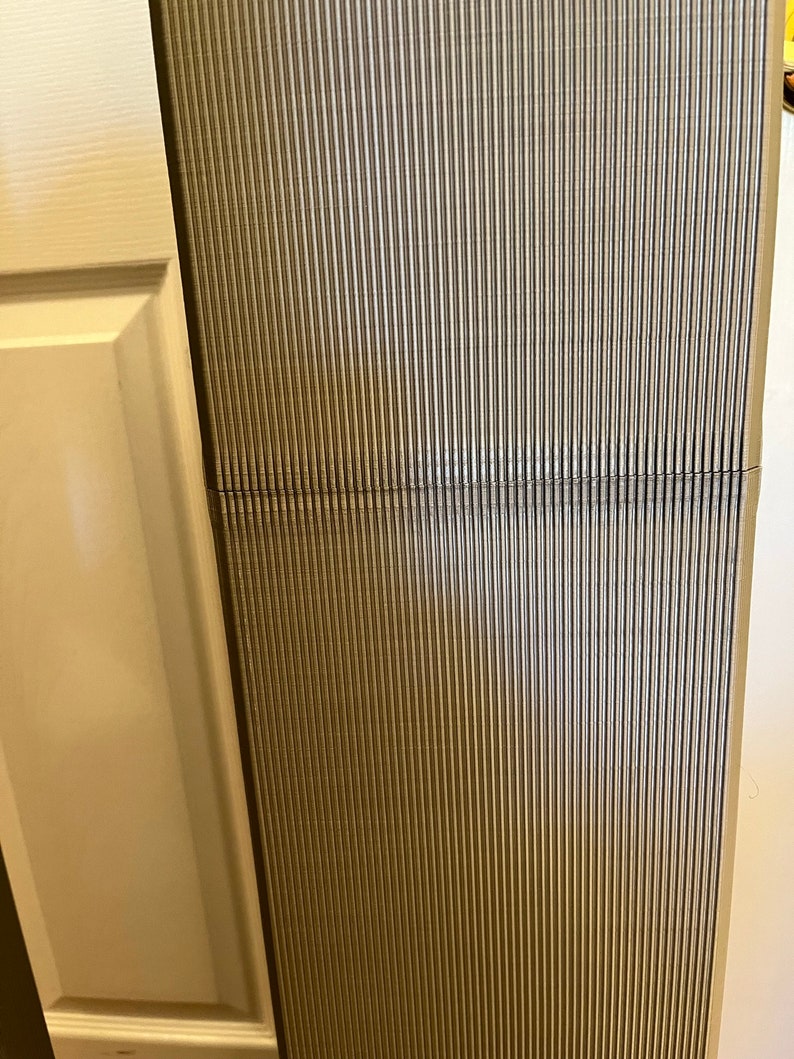 Pu&ograve; includere: Una sezione di una superficie verticale e scanalata in tonalit&agrave; di grigio e beige. Le scanalature sono parallele, creando un aspetto strutturato. Una linea orizzontale divide la superficie. Il bordo di un telaio di porta bianco &egrave; visibile a sinistra.