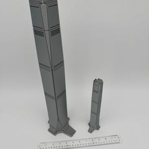 May include: Two gray, 3D-printed skyscraper models of different sizes. The larger model is approximately 12 inches tall, and the smaller one is about 6 inches. A ruler is placed at the bottom for scale, showing measurements in inches and centimeters.