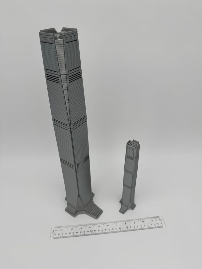 May include: Two gray, 3D-printed skyscraper models of different sizes. The larger model is approximately 12 inches tall, and the smaller one is about 6 inches. A ruler is placed at the bottom for scale, showing measurements in inches and centimeters.