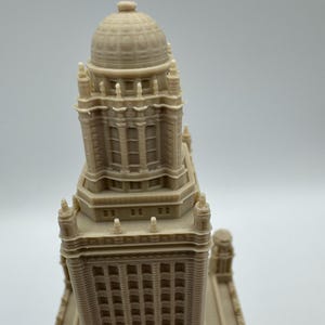 May include: A beige architectural model of a tall building with a domed top. The model has detailed windows and architectural features. The building's design includes multiple levels and decorative elements.
