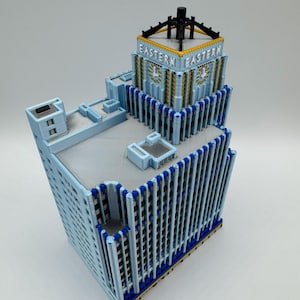May include: A light blue model of the Eastern building, featuring a detailed architectural design. The model has a rectangular shape with vertical lines and the word "EASTERN" on the top. Accents include gold and dark blue.
