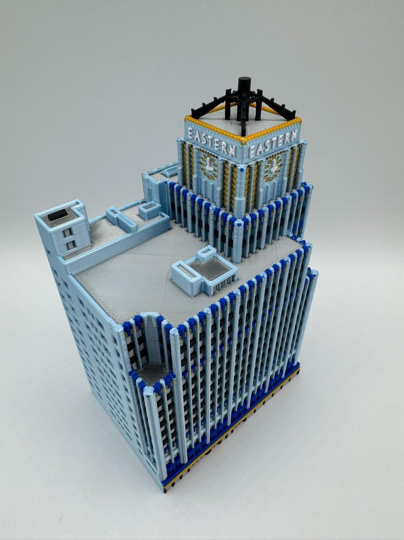 May include: A light blue model of the Eastern building, featuring a detailed architectural design. The model has a rectangular shape with vertical lines and the word "EASTERN" on the top. Accents include gold and dark blue.