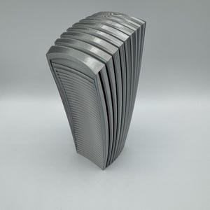 May include: A gray, abstract sculpture with a curved, layered design. The piece has a series of vertical, arched panels, creating a fan-like shape. The surface has a textured, ridged pattern, and the sculpture is set against a plain background.