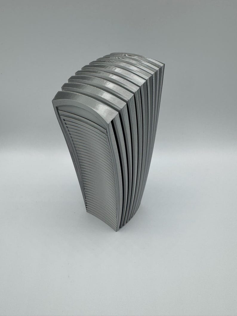 May include: A gray, abstract sculpture with a curved, layered design. The piece has a series of vertical, arched panels, creating a fan-like shape. The surface has a textured, ridged pattern, and the sculpture is set against a plain background.