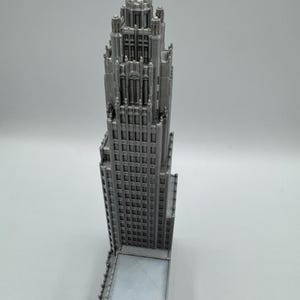 May include: A detailed, silver-colored model of a skyscraper. The building features a classic architectural design with numerous windows and intricate detailing on the upper levels. The model is a miniature representation of a tall building.