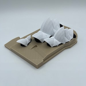 May include: A 3D printed model of the Sydney Opera House, featuring a white structure with a beige base. The model is made of plastic and is a popular souvenir for tourists.