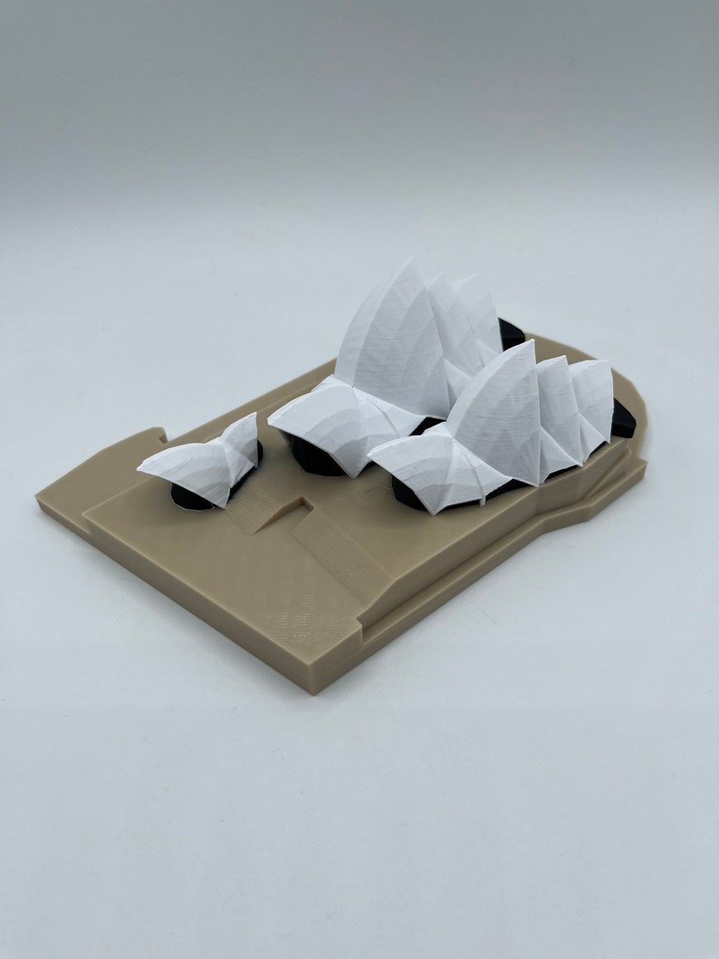 May include: A 3D printed model of the Sydney Opera House, featuring a white structure with a beige base. The model is made of plastic and is a popular souvenir for tourists.