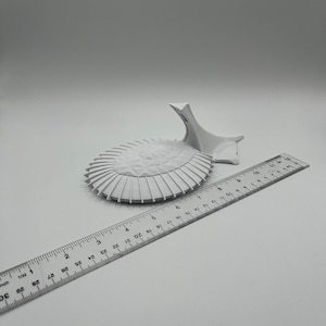 May include: A white 3D printed model of a building with a rounded roof and a tall, pointed spire. The model is sitting on a white surface next to a ruler.
