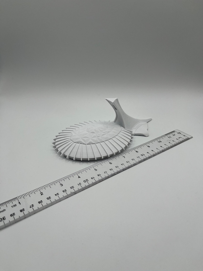 May include: A white 3D printed model of a building with a rounded roof and a tall, pointed spire. The model is sitting on a white surface next to a ruler.