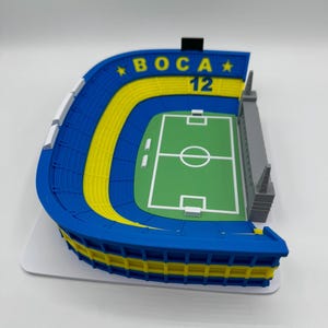 May include: A 3D-printed model of a football stadium in blue and yellow, with the text "BOCA 12" and a green pitch. The model shows a detailed design of a stadium, with seating areas and a playing field.