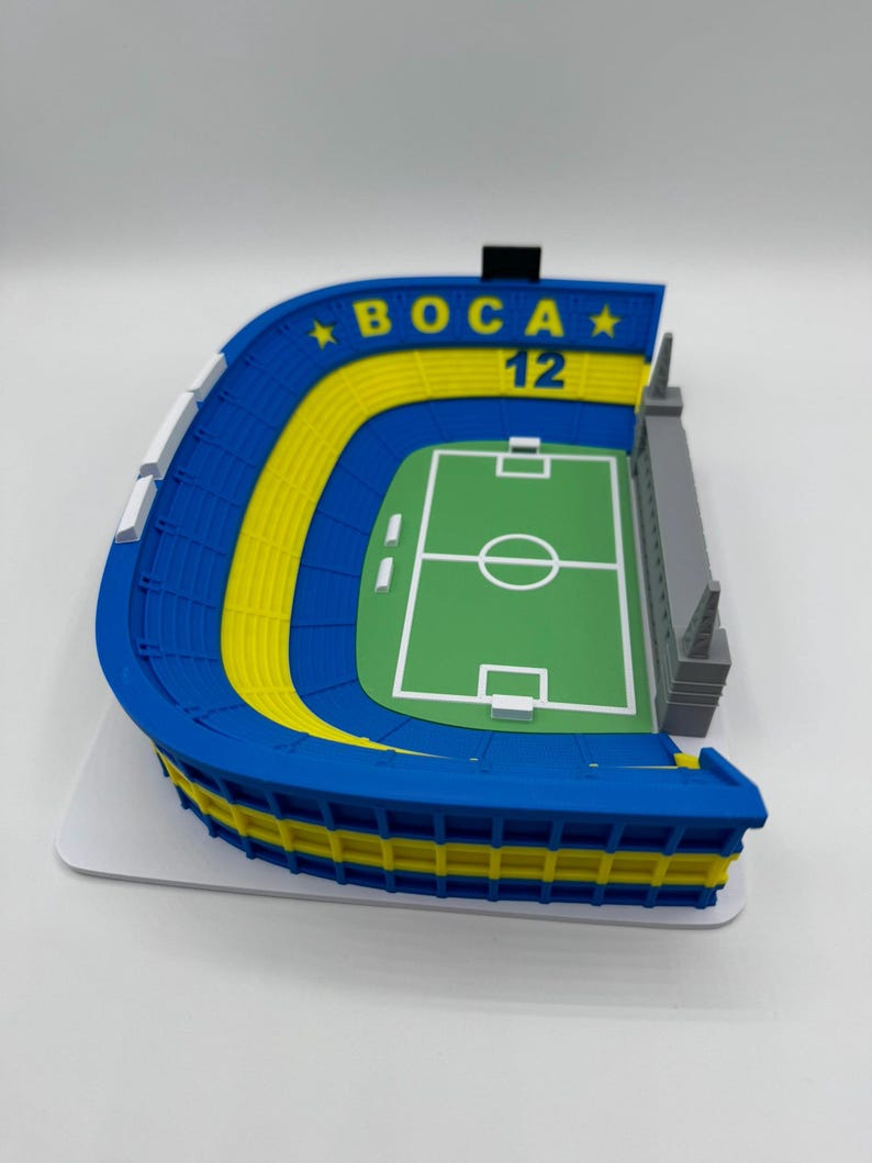 May include: A 3D-printed model of a football stadium in blue and yellow, with the text "BOCA 12" and a green pitch. The model shows a detailed design of a stadium, with seating areas and a playing field.