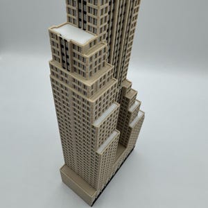 May include: A beige miniature replica of a skyscraper, featuring multiple rectangular sections with numerous windows. The building has a flat roof and a dark base. The model is a detailed representation of an iconic architectural structure.