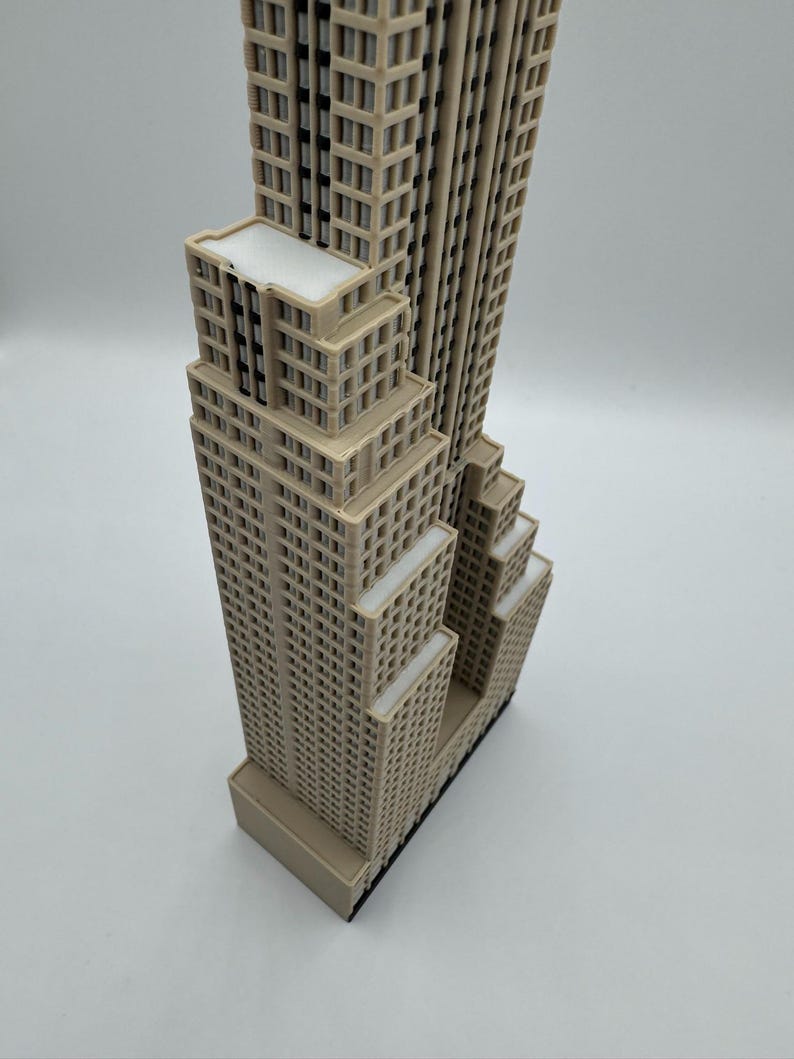 May include: A beige miniature replica of a skyscraper, featuring multiple rectangular sections with numerous windows. The building has a flat roof and a dark base. The model is a detailed representation of an iconic architectural structure.