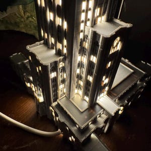 May include: A gray 3D-printed architectural model of a skyscraper featuring multiple tiered levels and illuminated windows that emit a warm light. The structure has a textured surface and is connected to a white power cord at the base.