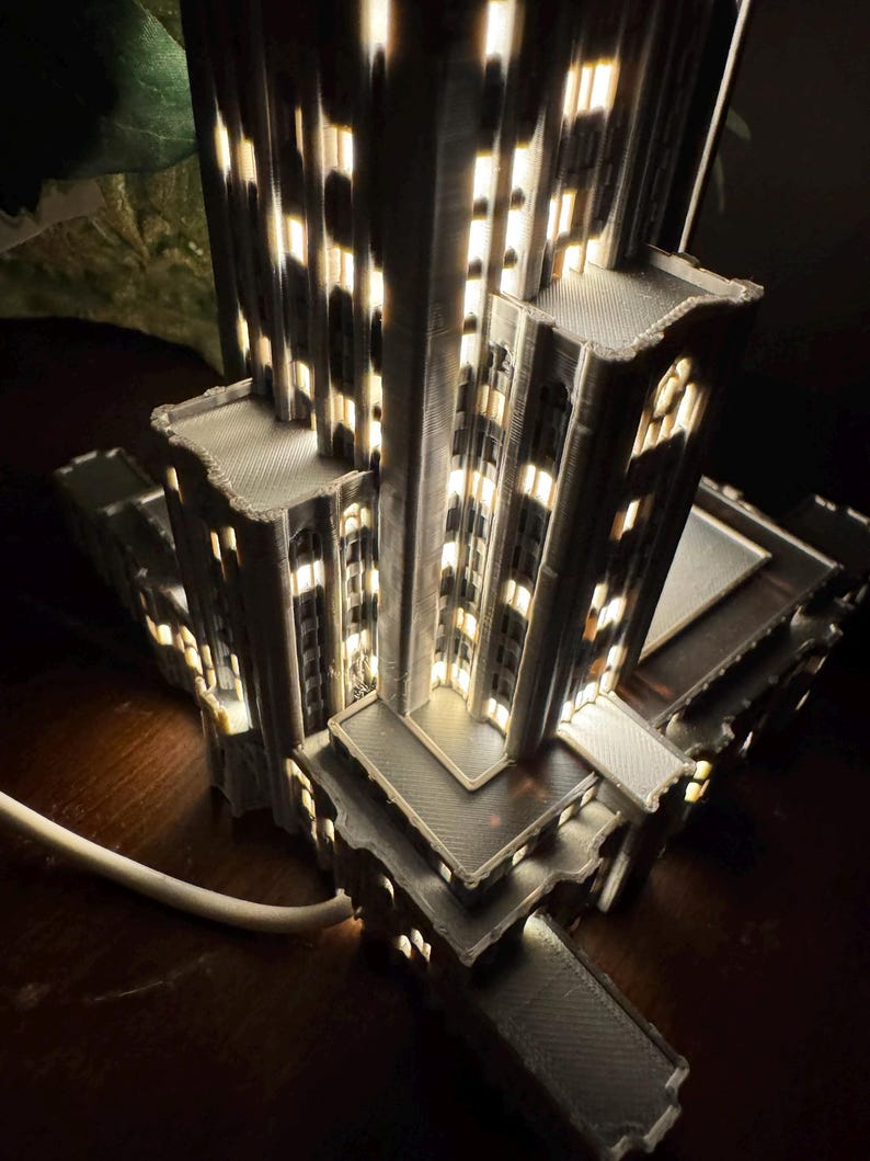 May include: A gray 3D-printed architectural model of a skyscraper featuring multiple tiered levels and illuminated windows that emit a warm light. The structure has a textured surface and is connected to a white power cord at the base.