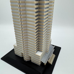 May include: A 3D-printed architectural model of a skyscraper. The building features a layered design with alternating beige, white, and gray sections. It sits on a black rectangular base, showcasing intricate details and a modern aesthetic.