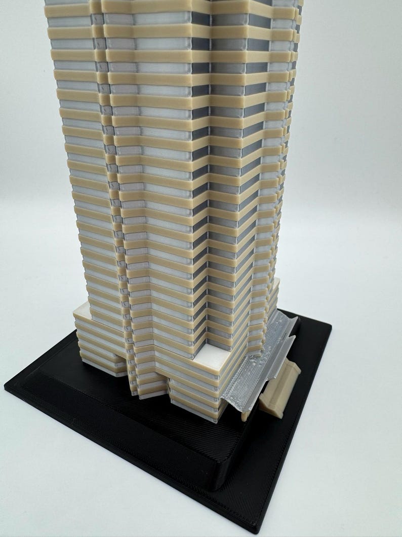 May include: A 3D-printed architectural model of a skyscraper. The building features a layered design with alternating beige, white, and gray sections. It sits on a black rectangular base, showcasing intricate details and a modern aesthetic.