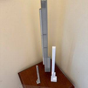 Op de afbeelding: Drie 3D-geprinte modellen van wolkenkrabbers, &eacute;&eacute;n is grijs en de andere twee zijn wit. De modellen staan op een houten tafel met een liniaal.