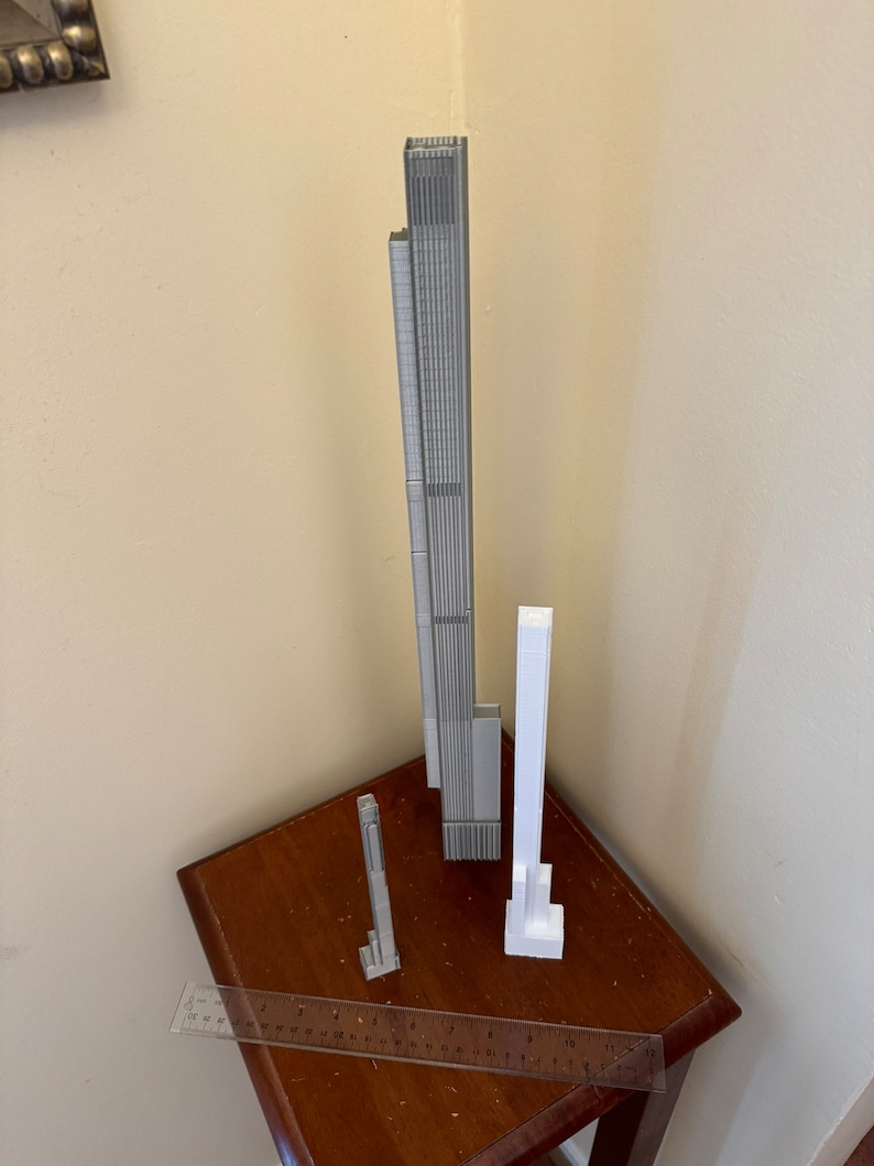 Op de afbeelding: Drie 3D-geprinte modellen van wolkenkrabbers, &eacute;&eacute;n is grijs en de andere twee zijn wit. De modellen staan op een houten tafel met een liniaal.