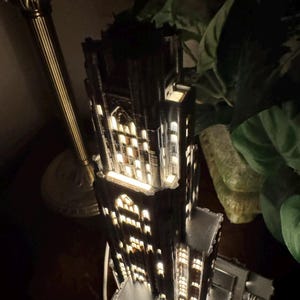 May include: A dark-colored, multi-tiered architectural model of a skyscraper featuring intricate gothic-style window cutouts. The structure is illuminated from within, casting a warm glow through the numerous rectangular and arched openings.