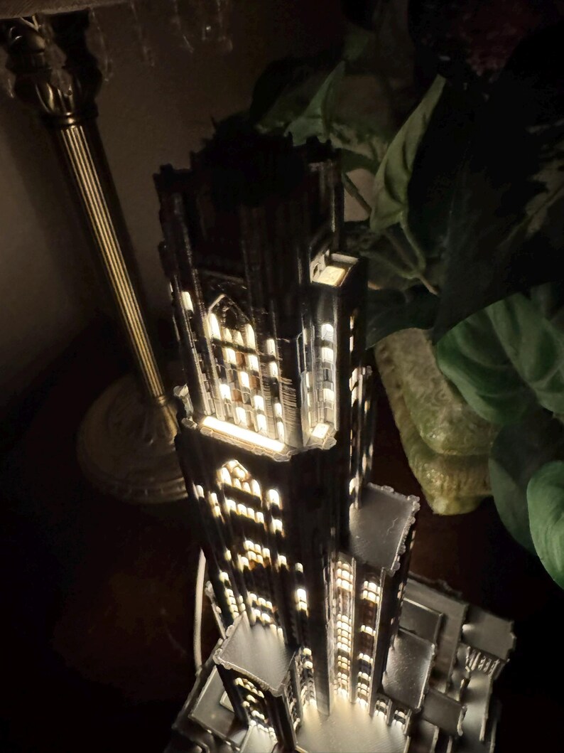May include: A dark-colored, multi-tiered architectural model of a skyscraper featuring intricate gothic-style window cutouts. The structure is illuminated from within, casting a warm glow through the numerous rectangular and arched openings.