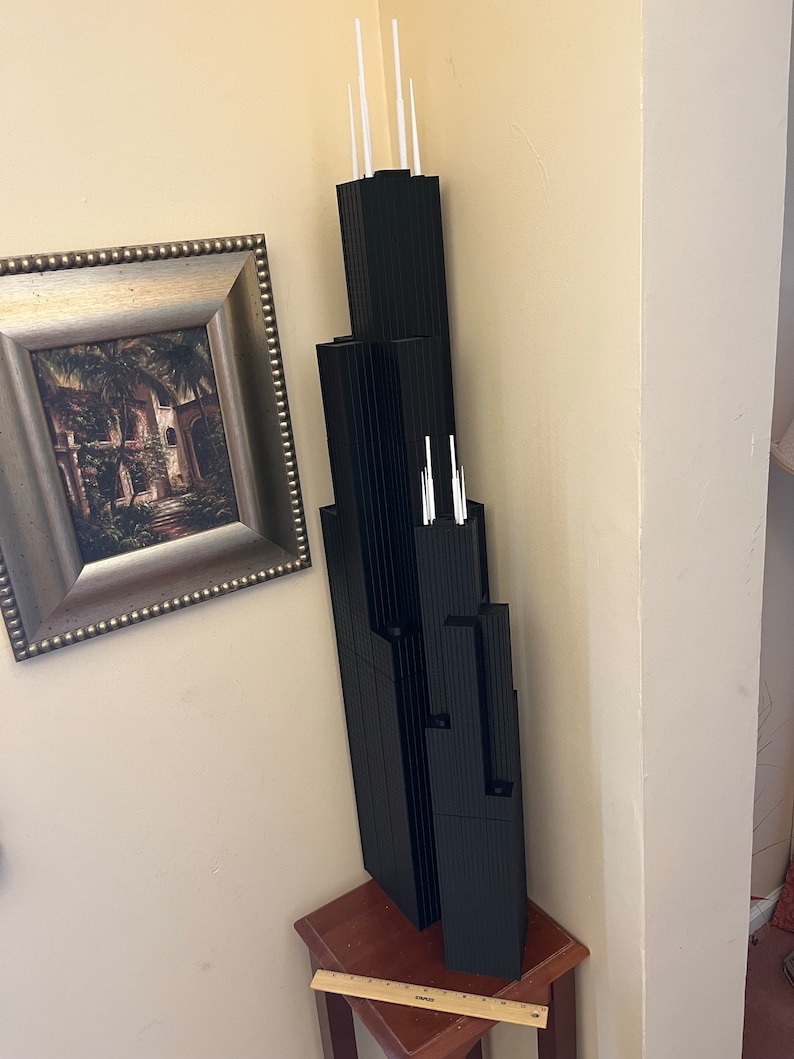 May include: A black model of the Willis Tower, formerly known as the Sears Tower, with white spires on top. The model is sitting on a wooden table.