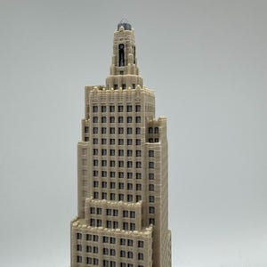 May include: A detailed miniature model of a beige skyscraper with a tiered design. The building features numerous windows and architectural details, with a spire at the top. The model is set against a plain background.