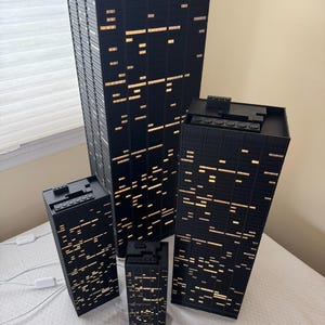 Peut inclure: Ensemble de quatre maquettes de gratte-ciel noirs avec des fen&ecirc;tres &eacute;clair&eacute;es. Les b&acirc;timents sont de tailles diff&eacute;rentes, le plus grand mesurant environ 46 cm de haut. Les fen&ecirc;tres &eacute;mettent une lumi&egrave;re chaude, simulant un paysage urbain nocturne.