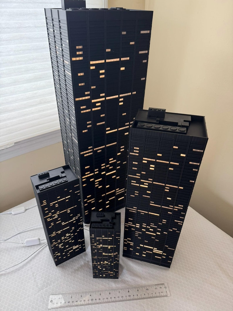 Peut inclure: Ensemble de quatre maquettes de gratte-ciel noirs avec des fen&ecirc;tres &eacute;clair&eacute;es. Les b&acirc;timents sont de tailles diff&eacute;rentes, le plus grand mesurant environ 46 cm de haut. Les fen&ecirc;tres &eacute;mettent une lumi&egrave;re chaude, simulant un paysage urbain nocturne.