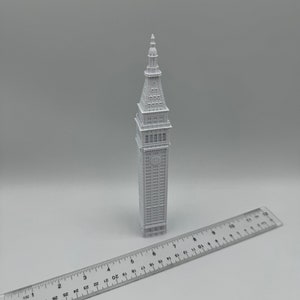 May include: A white 3D printed model of a tall building with a pointed roof. The model is about 7 inches tall and is sitting on a white surface next to a ruler.