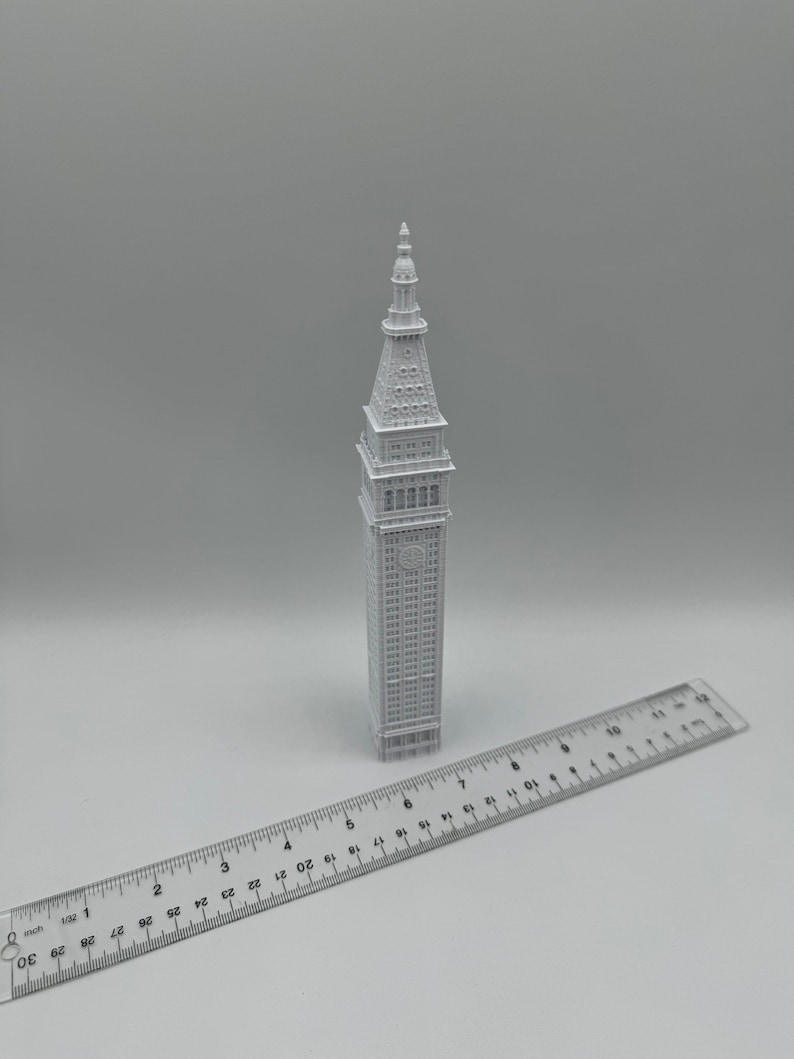 May include: A white 3D printed model of a tall building with a pointed roof. The model is about 7 inches tall and is sitting on a white surface next to a ruler.