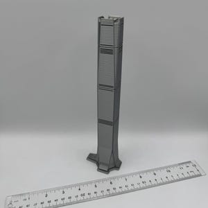 May include: A gray miniature skyscraper model with a detailed facade. A ruler is placed next to the model for scale. The building has horizontal lines and a unique base. The model is approximately 10 inches tall.