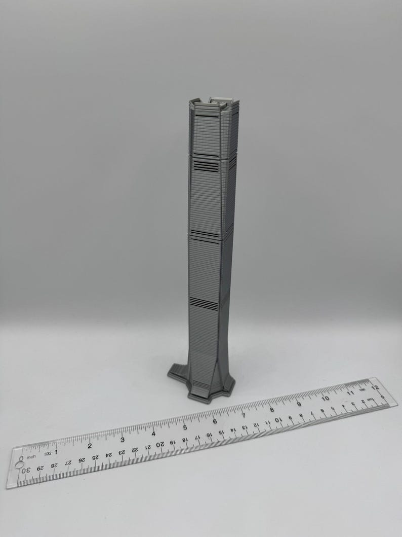 May include: A gray miniature skyscraper model with a detailed facade. A ruler is placed next to the model for scale. The building has horizontal lines and a unique base. The model is approximately 10 inches tall.