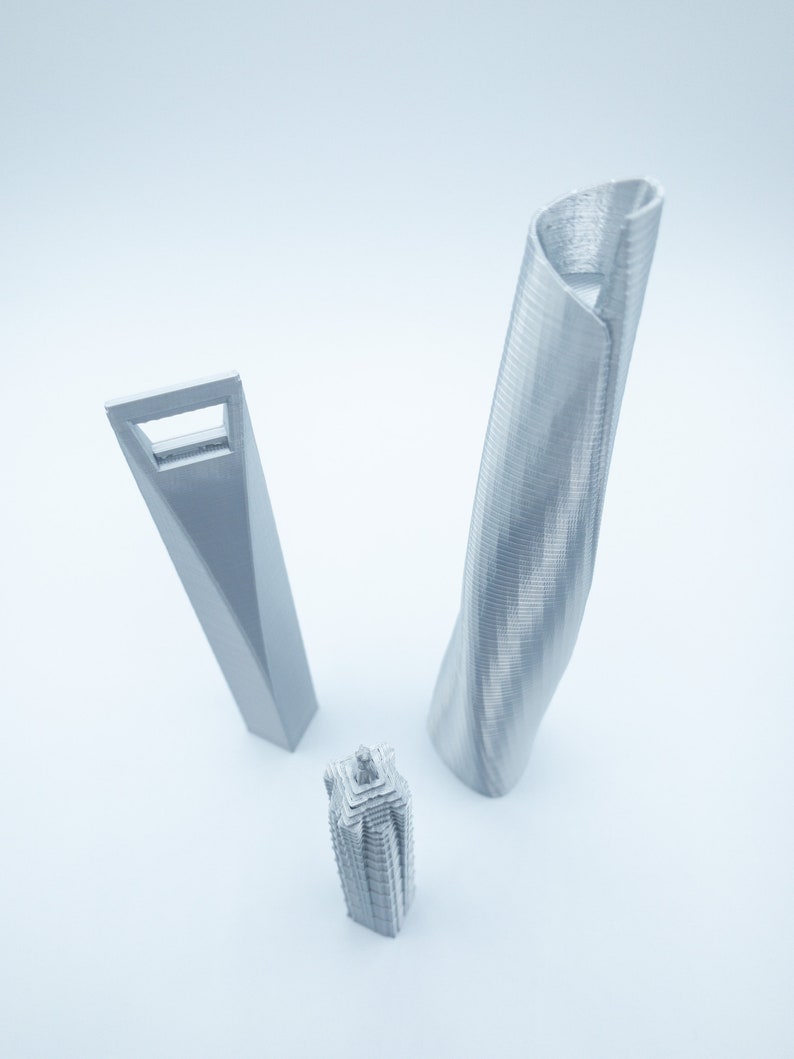 May include: Three silver 3D printed models of skyscrapers. The tallest model is a twisted tower, the middle model is a rectangular tower with a window, and the shortest model is a square tower with a pointed top.
