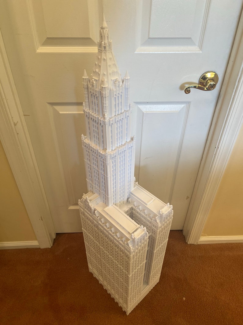 May include: A white 3D printed model of a tall skyscraper with a pointed roof and intricate details.