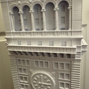 Op de afbeelding: Een wit 3D-geprint model van een hoog gebouw met een klok op de voorgevel. Het gebouw heeft veel ramen en een decoratieve kroonlijst.